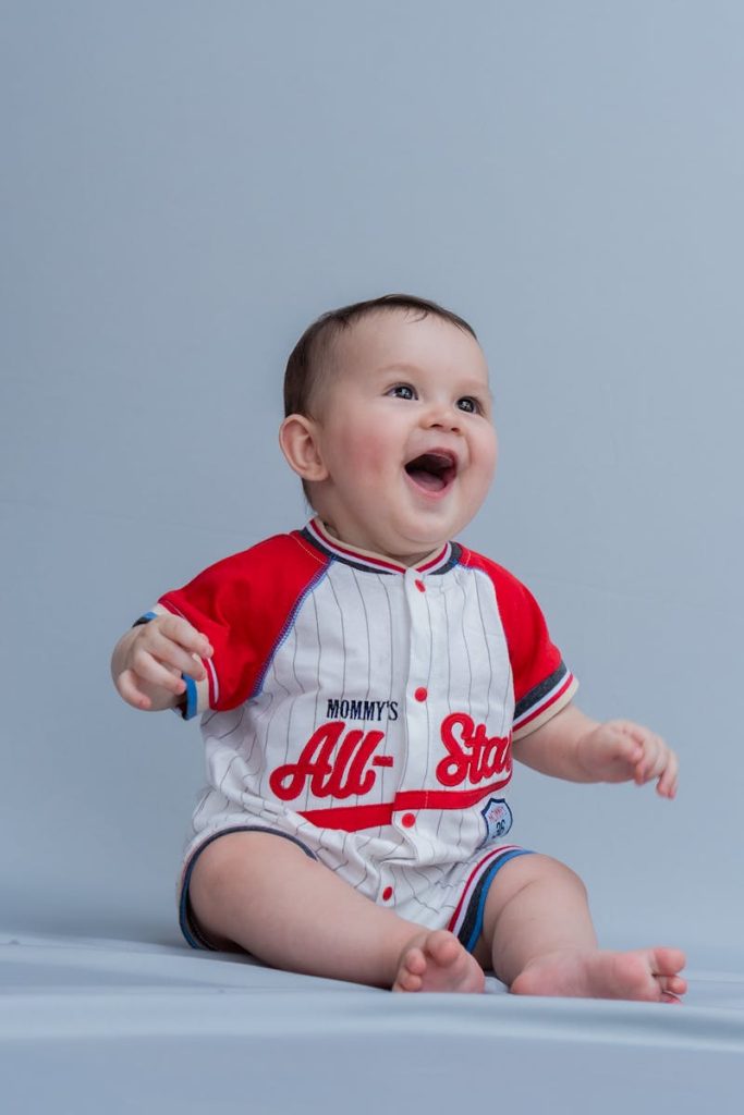 Smiling baby wearing a baseball-themed romper, sitting on a light blue background.