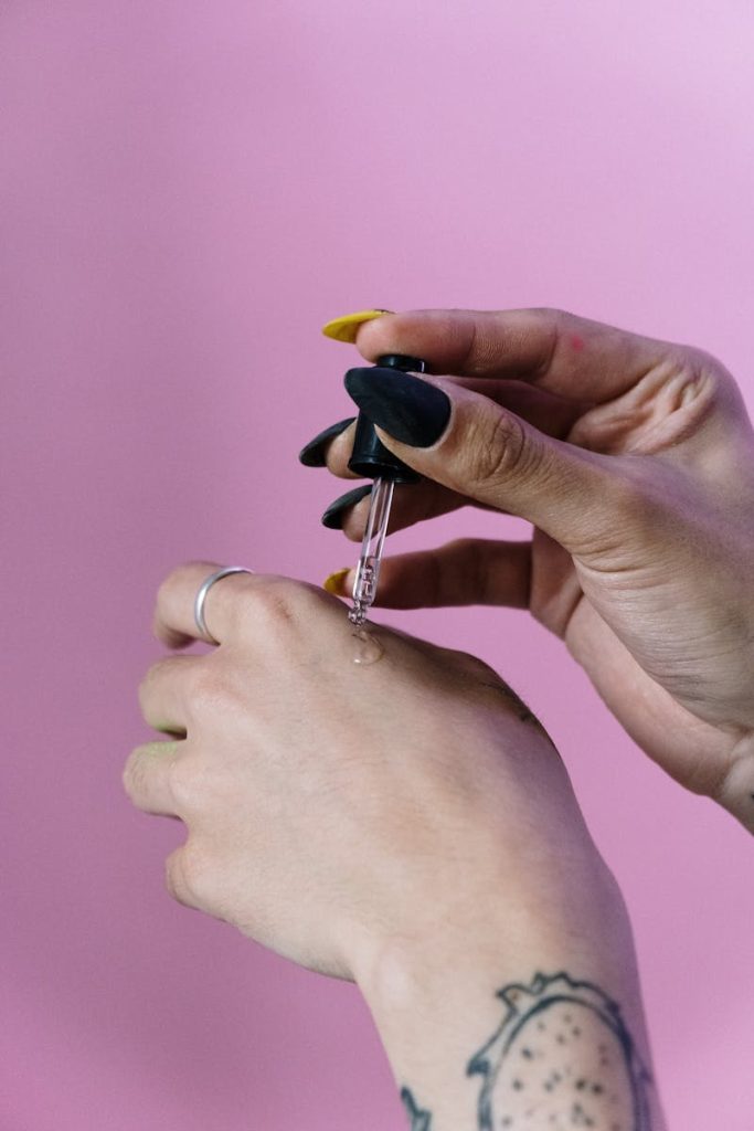 A close-up of a woman's hand applying serum with a dropper against a pink background.
